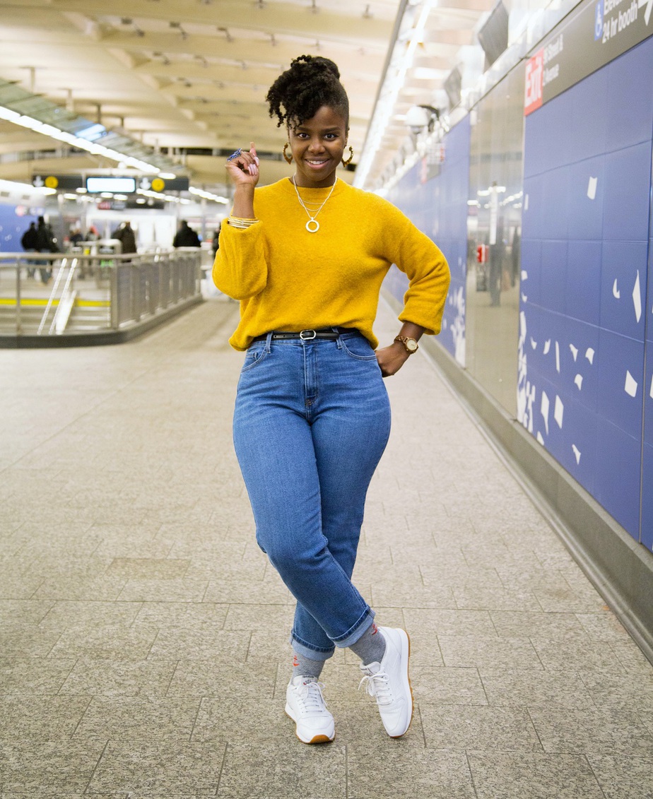Toia Barry standing in a train station wearing yellow cozy fall sweater, legs crossed