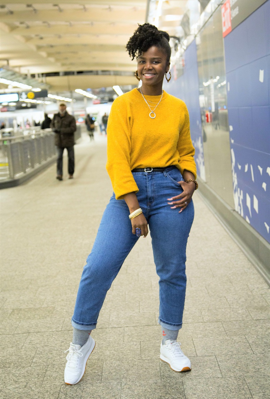 black woman wearing a yellow cozy fall sweater, posed in a NYC train station with one hand in jeans pocket.